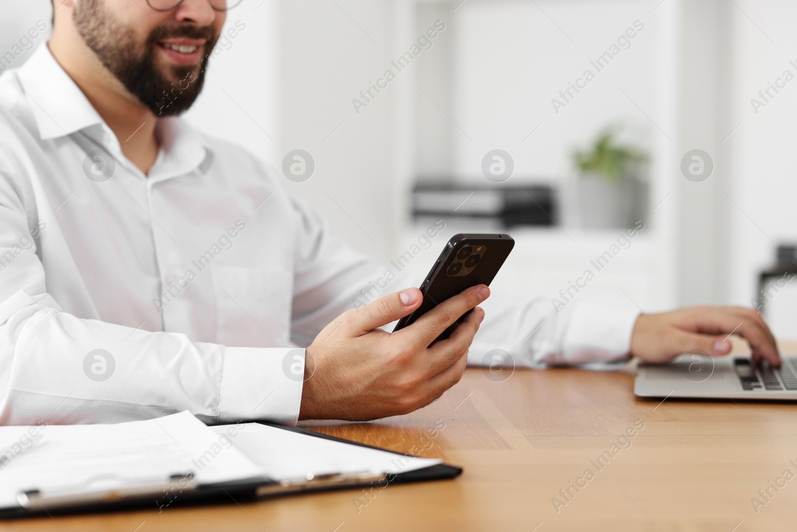 Photo of Young man using smartphone while working with laptop at wooden table in office, closeup