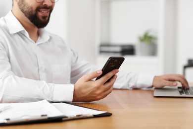 Photo of Young man using smartphone while working with laptop at wooden table in office, closeup