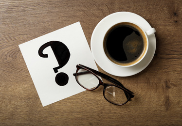 Photo of Note with question mark, eyeglasses and cup of coffee on wooden table, flat lay
