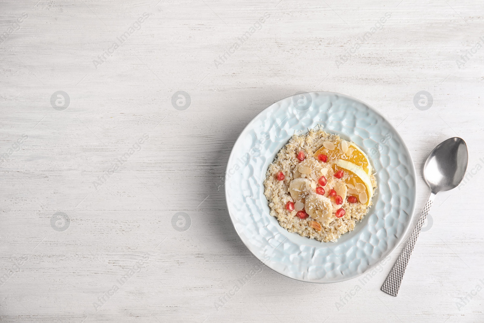 Photo of Plate of quinoa porridge with orange, banana and pomegranate seeds near spoon on white wooden background, top view. Space for text