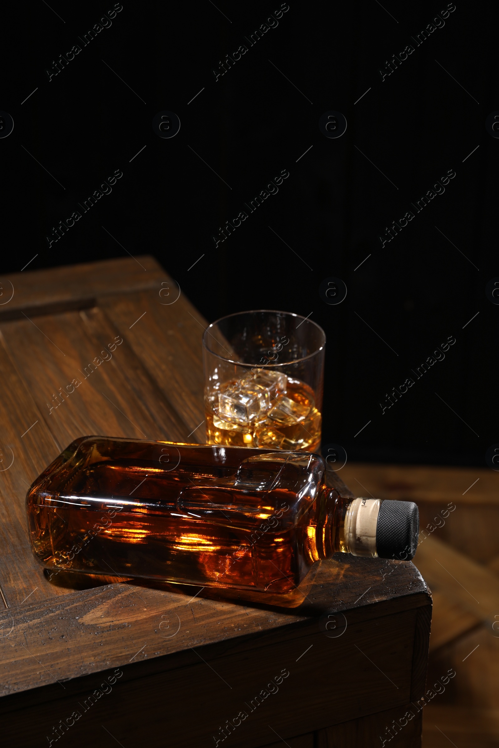 Photo of Whiskey with ice cubes in glass and bottle on wooden crate against black background