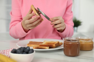 Photo of Woman spreading tasty nut butter onto toast at white marble table, closeup