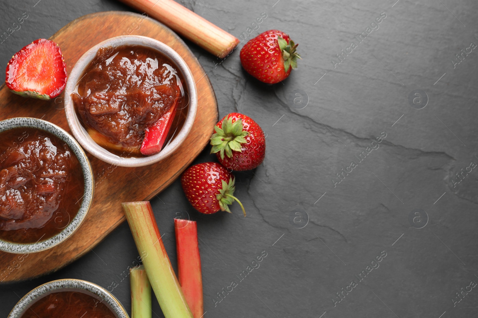 Photo of Tasty rhubarb jam in bowls, stems and strawberries on dark textured table, flat lay. Space for text