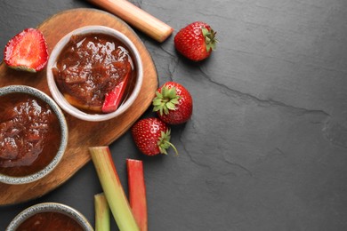 Photo of Tasty rhubarb jam in bowls, stems and strawberries on dark textured table, flat lay. Space for text