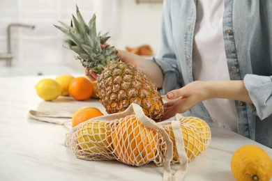 Photo of Woman taking pineapple out from string bag at light marble table, closeup