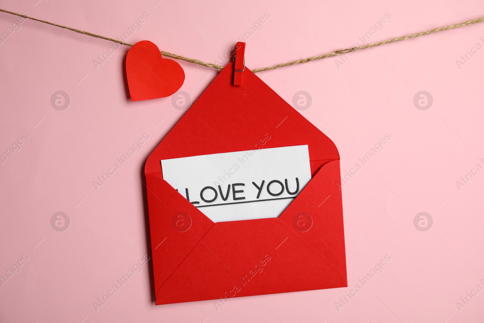 Photo of Red envelope with Love You card and paper heart hanging on twine against pink background