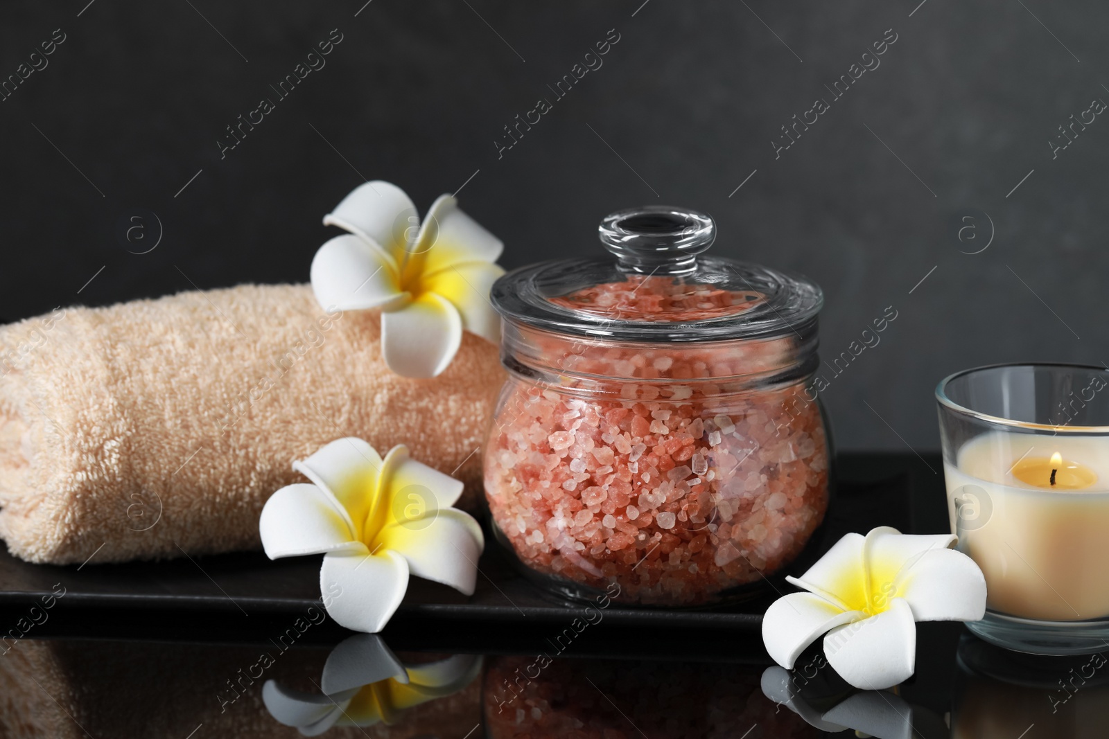 Photo of Glass jar with natural sea salt and beautiful flowers on mirror surface