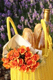 Yellow wicker bag with beautiful roses, bottle of wine and baguettes in lavender field, closeup