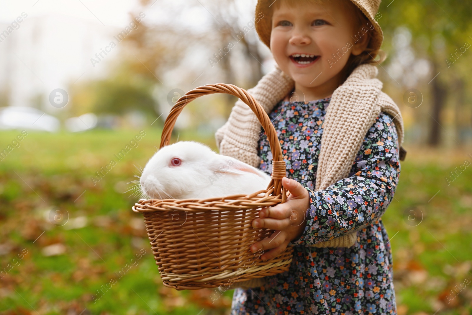 Photo of Happy girl holding basket with cute white rabbit in autumn park, focus on pet