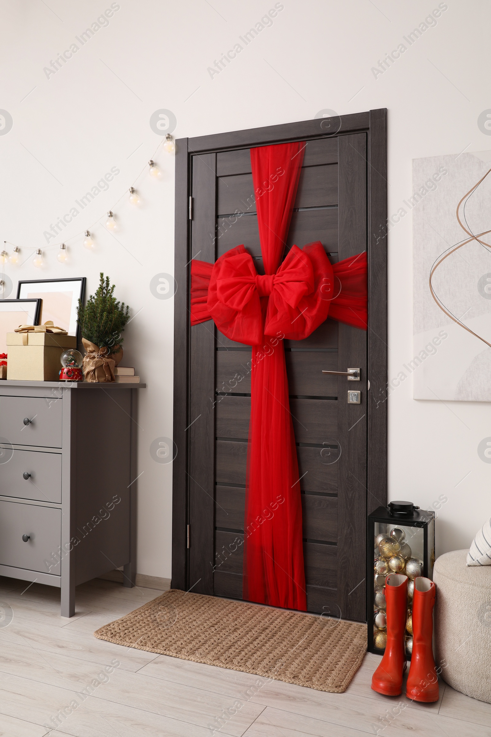 Photo of Wooden door with beautiful red bow near chest of drawers and container with baubles indoors. Christmas decoration