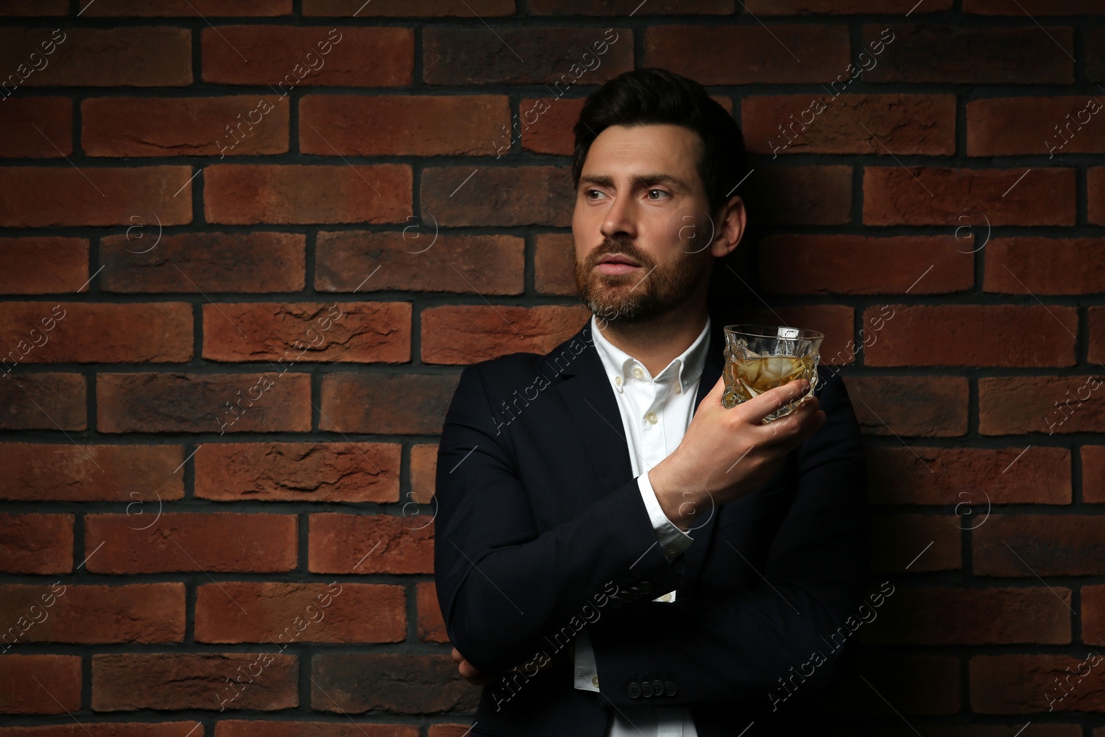 Photo of Handsome man in formal suit holding glass of whiskey near red brick wall. Space for text