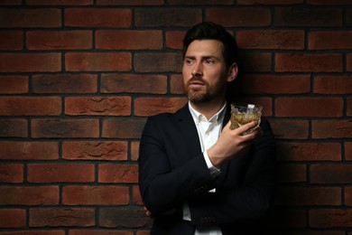 Handsome man in formal suit holding glass of whiskey near red brick wall. Space for text