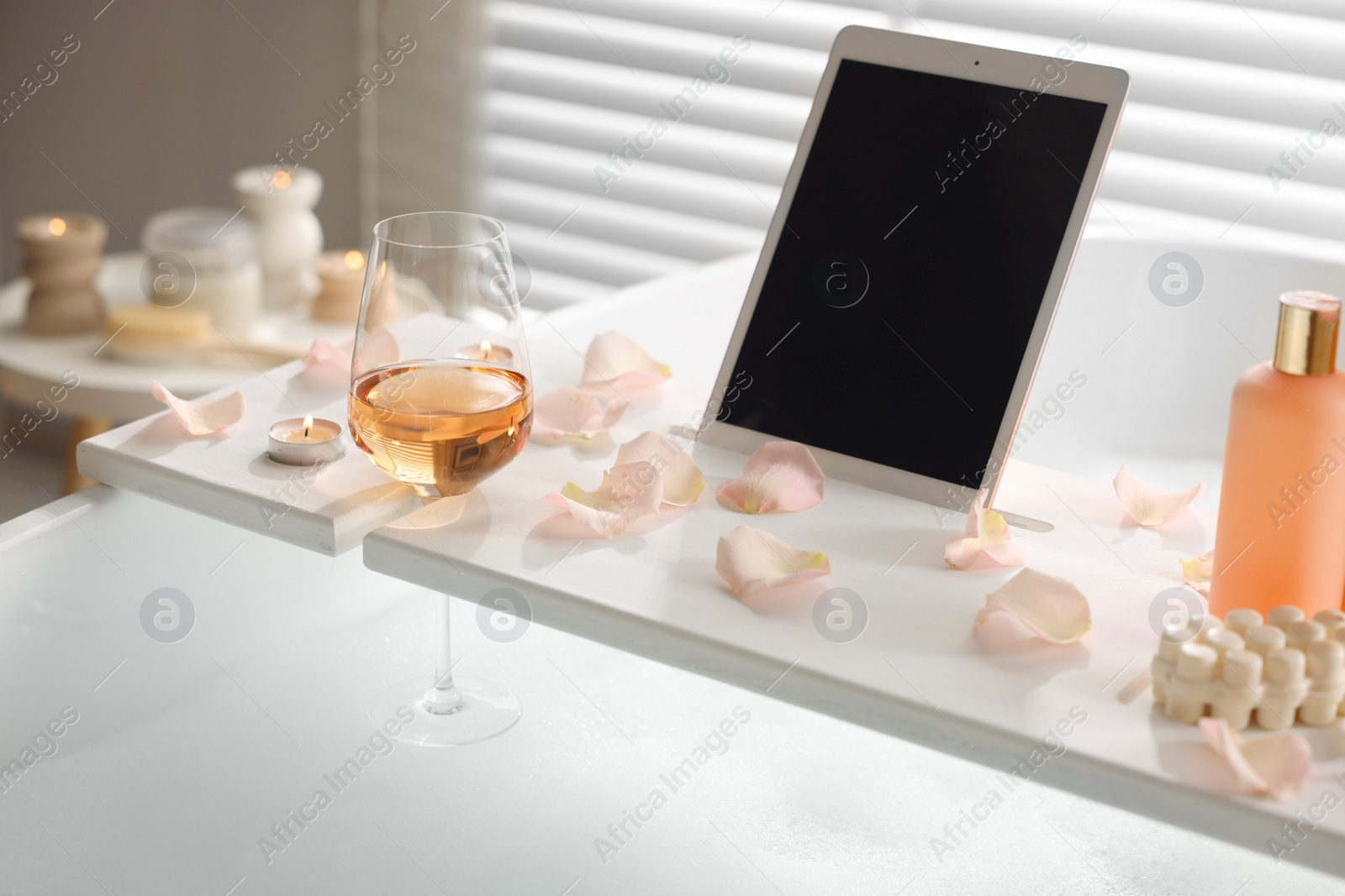 Photo of Wooden tray with tablet, wine, toiletries and flower petals on bathtub in bathroom