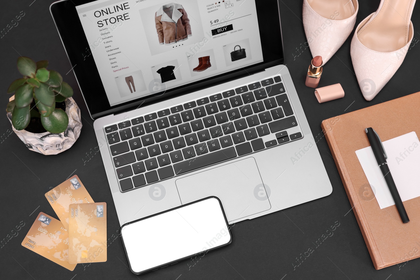 Photo of Online store website on laptop screen. Computer, smartphone, credit cards, women's shoes, stationery and lipstick on black background, above view