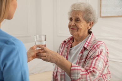 Photo of Young caregiver giving water to senior woman in room. Home care service