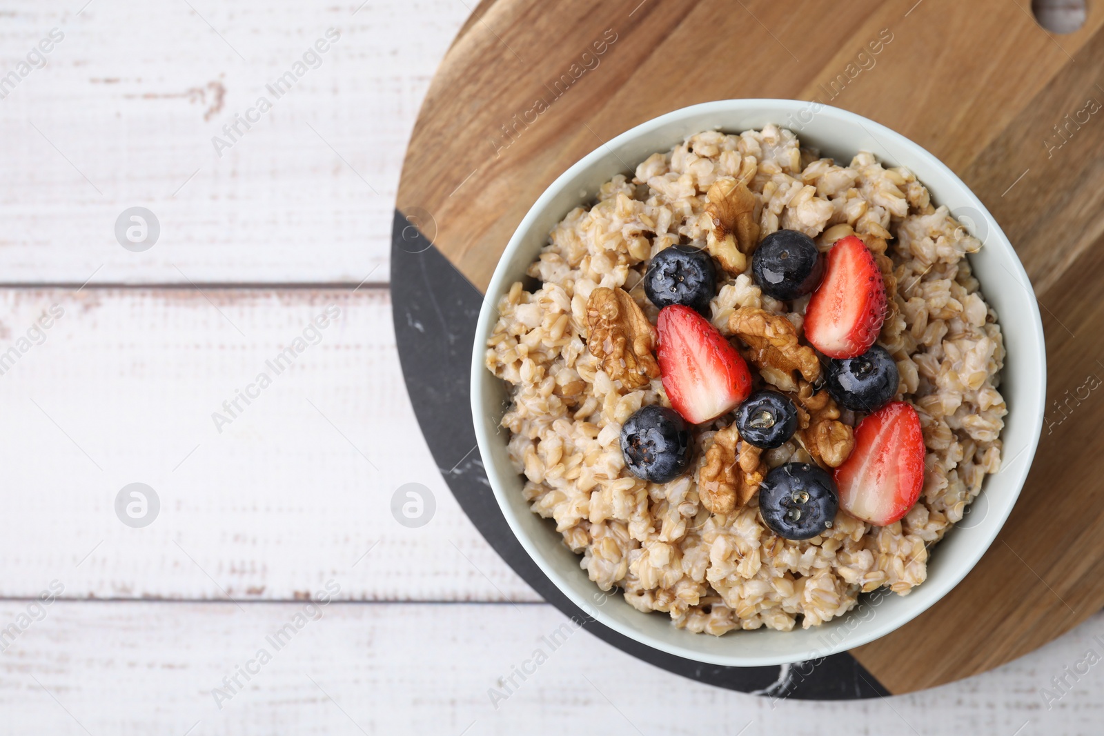 Photo of Tasty oatmeal with strawberries, blueberries and walnuts in bowl on white wooden table, top view