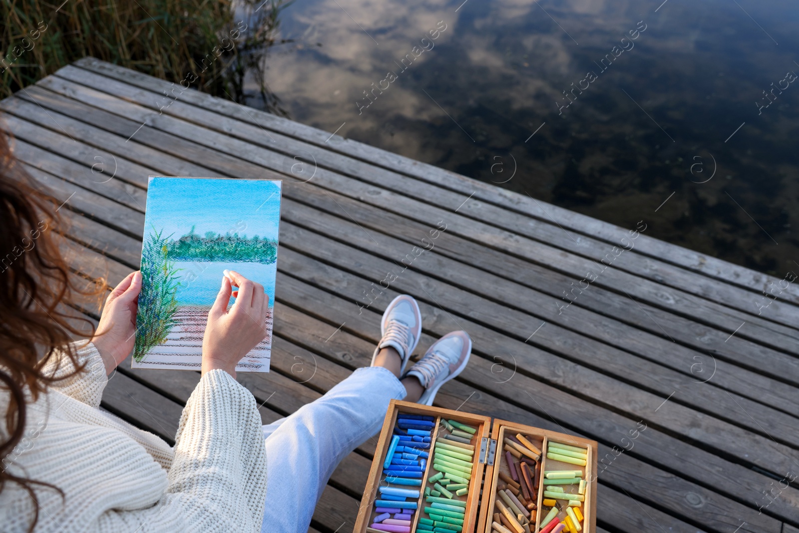 Photo of Woman drawing with soft pastels on wooden pier near river, closeup