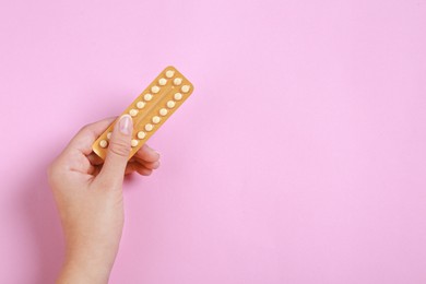 Woman holding blister with birth control pills on pink background, top view. Space for text