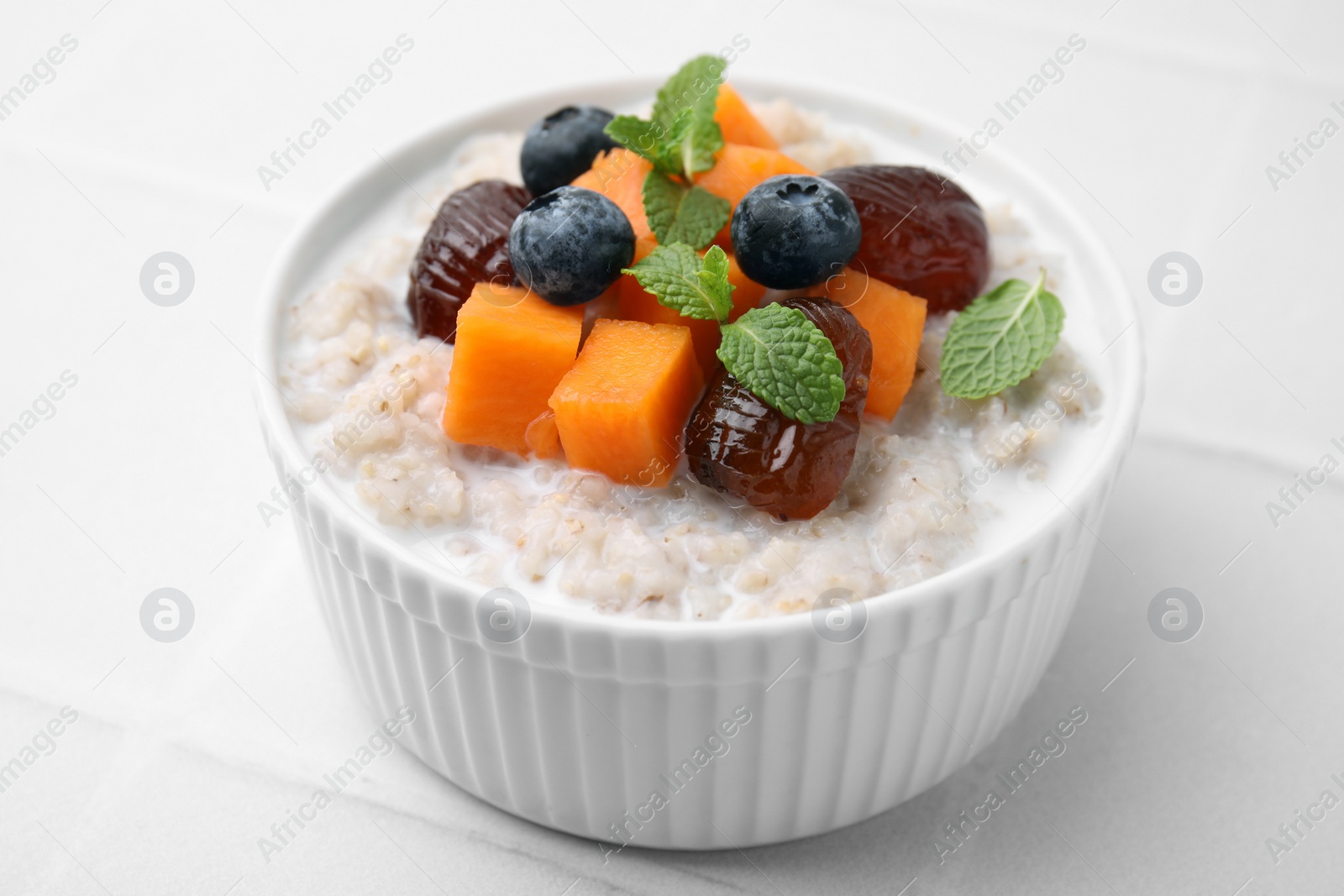Photo of Delicious barley porridge with blueberries, pumpkin, dates and mint in bowl on white tiled table, closeup