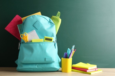 Photo of Backpack full of different school stationery on table near blank chalkboard. Space for text
