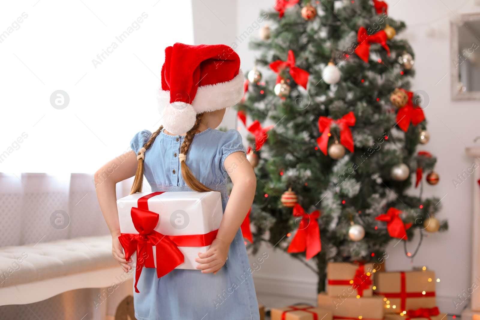 Photo of Cute little child in Santa hat hiding Christmas gift box behind back at home