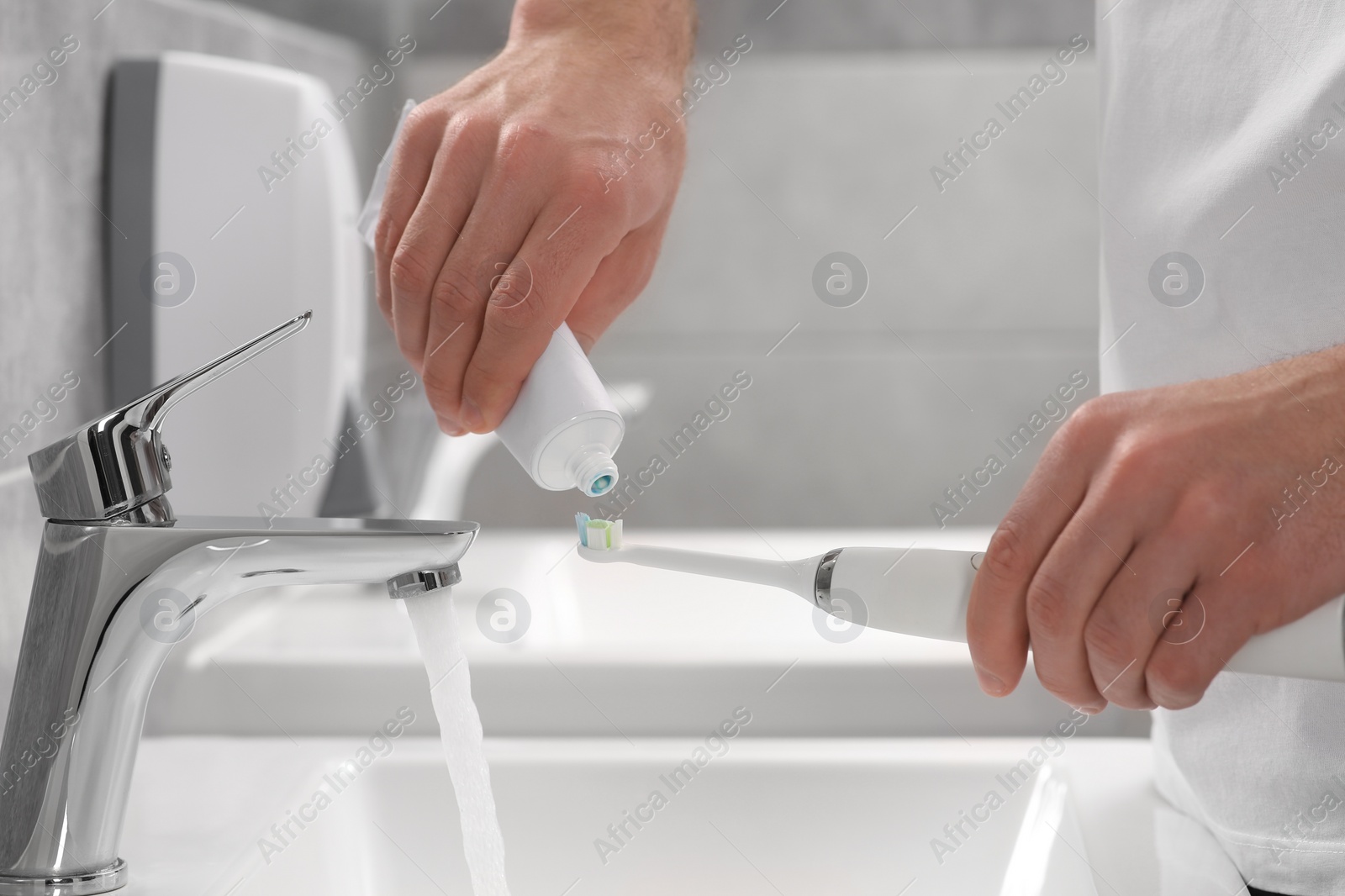 Photo of Man squeezing toothpaste from tube onto electric toothbrush in bathroom, closeup