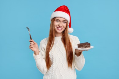 Young woman in Santa hat with piece of tasty cake on light blue background