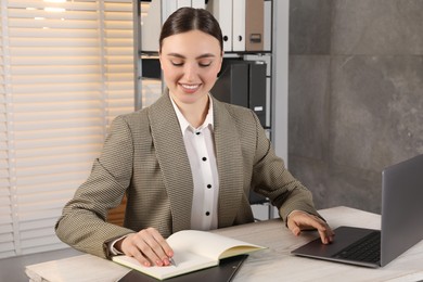 Photo of Happy woman taking notes while using laptop at light wooden table in office