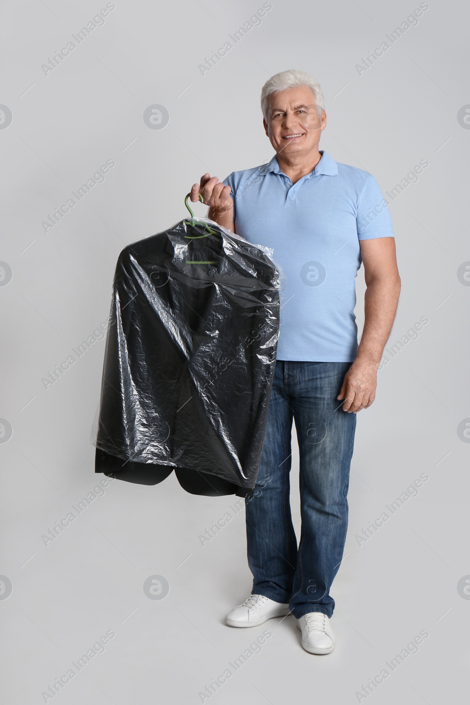 Photo of Senior man holding hanger with jacket in plastic bag on light grey background. Dry-cleaning service