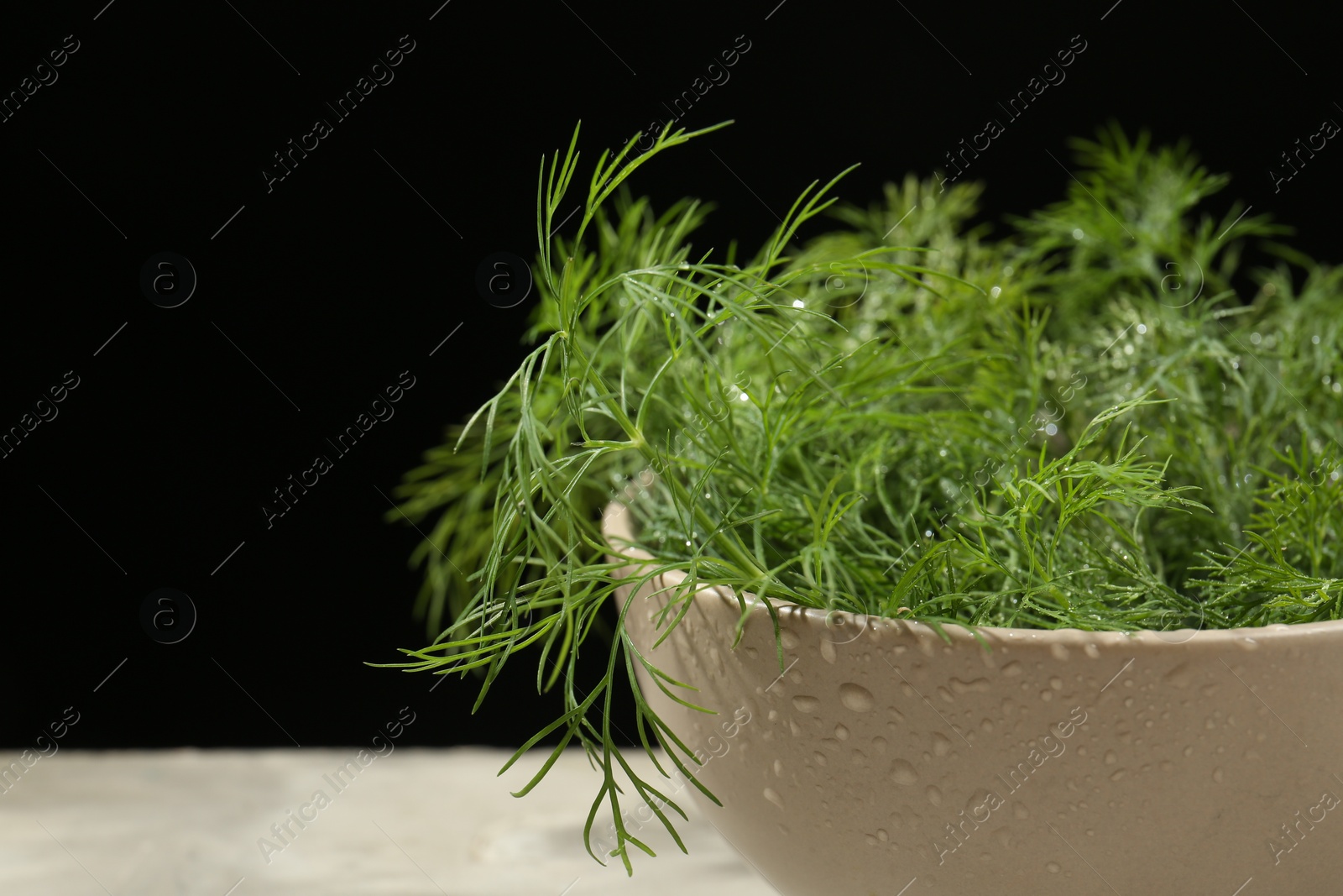 Photo of Bowl of fresh green dill with water drops on light grey table against black background, closeup. Space for text