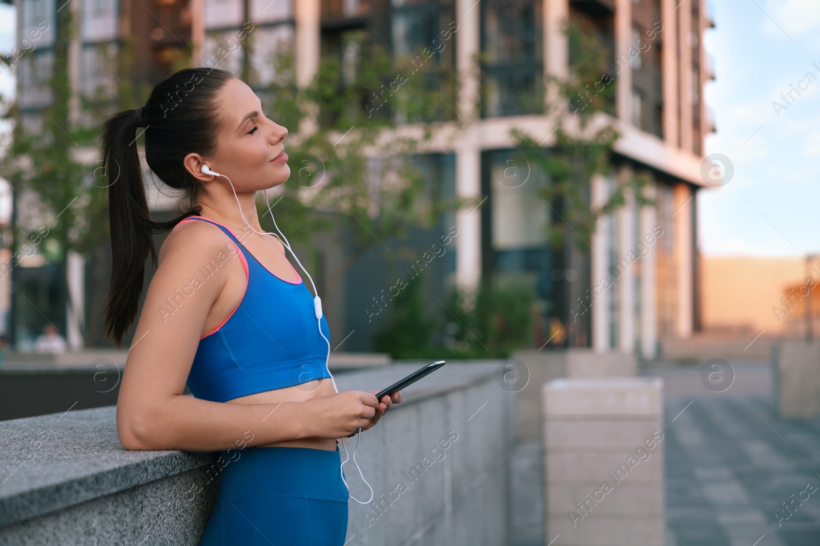 Photo of Beautiful woman in stylish sportswear listening music on city street, space for text