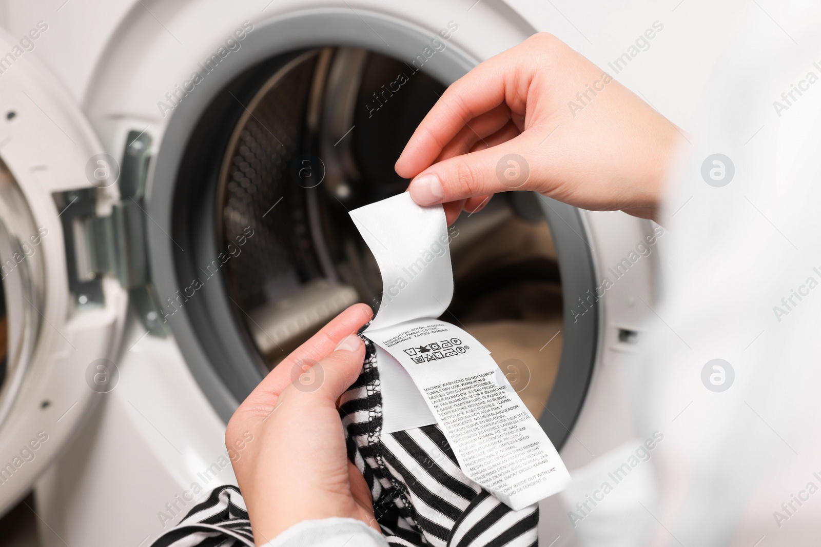 Photo of Woman reading clothing label with care symbols and material content on striped shirt near washing machine, closeup