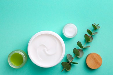 Moisturizing cream in open jars and eucalyptus branch on turquoise background, flat lay. Body care products