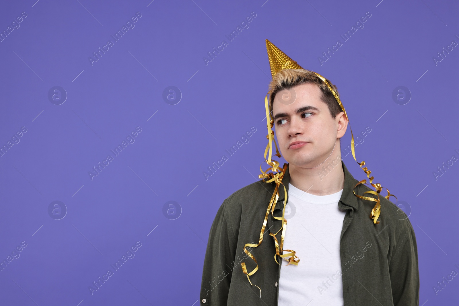 Photo of Sad young man with party hat on purple background, space for text
