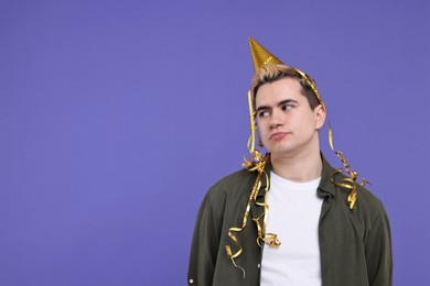 Sad young man with party hat on purple background, space for text