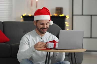 Photo of Celebrating Christmas online with exchanged by mail presents. Smiling man in Santa hat opening gift box during video call on laptop at home