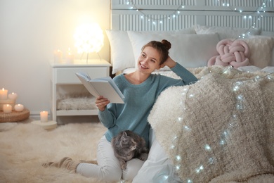 Photo of Young woman with cute cat reading book at home. Cozy winter