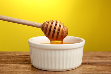 Pouring honey from dipper into bowl at wooden table against yellow background, closeup