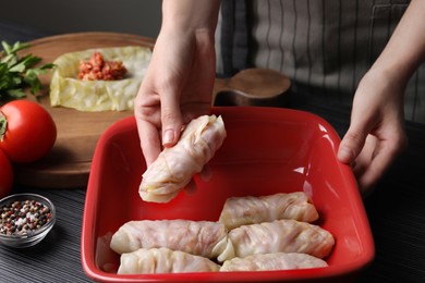 Woman putting uncooked stuffed cabbage roll into baking dish at black table, closeup