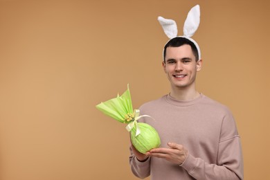 Easter celebration. Handsome young man with bunny ears holding wrapped gift on beige background. Space for text