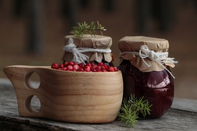 Photo of Tasty lingonberry jam in jars and cup with red berries on wooden table outdoors, closeup