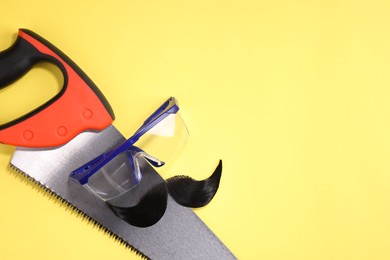 Man's face made of artificial mustache, safety glasses and hand saw on yellow background, top view. Space for text