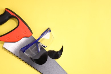 Photo of Man's face made of artificial mustache, safety glasses and hand saw on yellow background, top view. Space for text