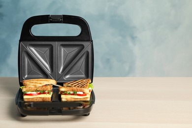 Modern grill maker with sandwiches on light wooden table, space for text