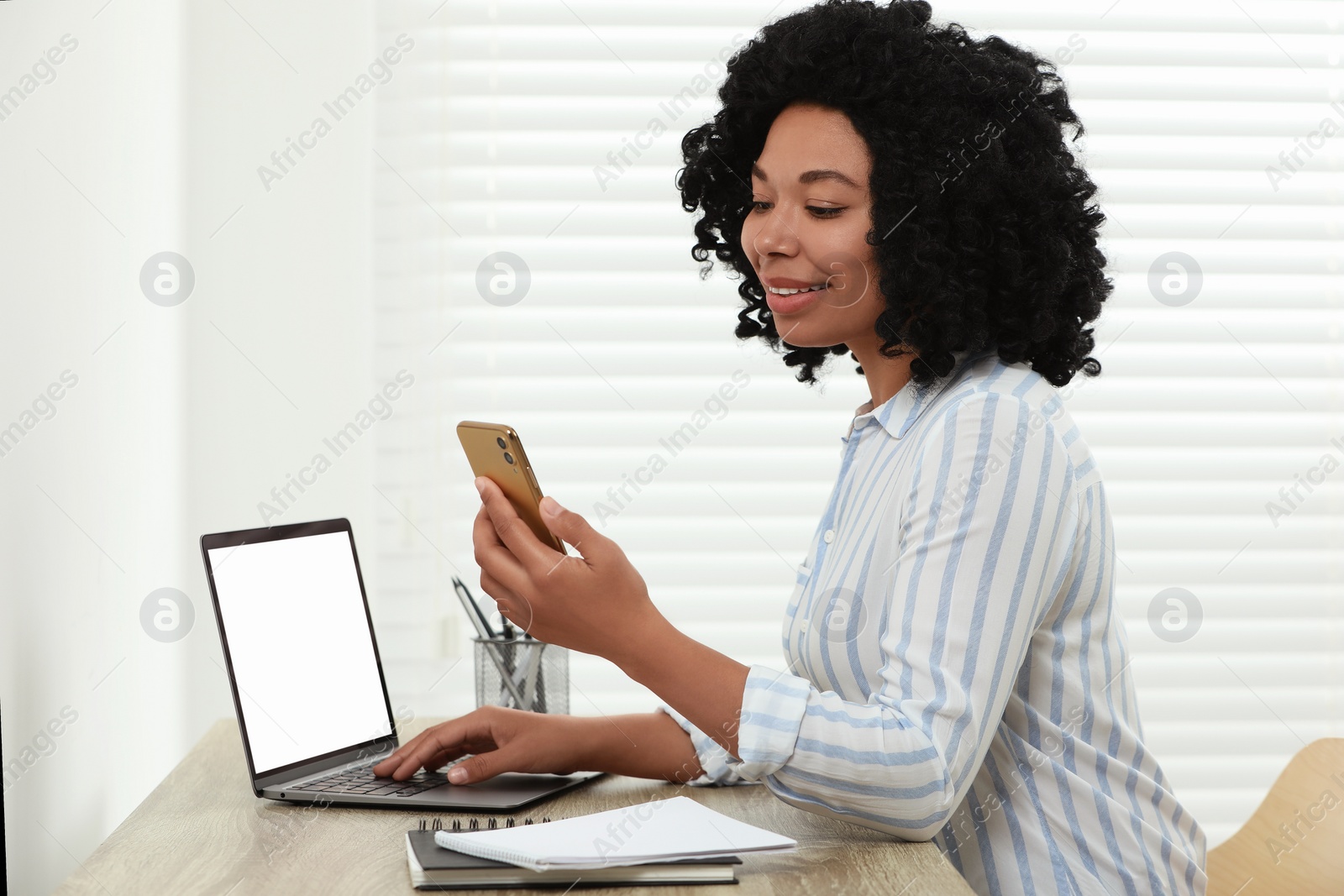 Photo of Happy young woman with smartphone using laptop at wooden desk indoors