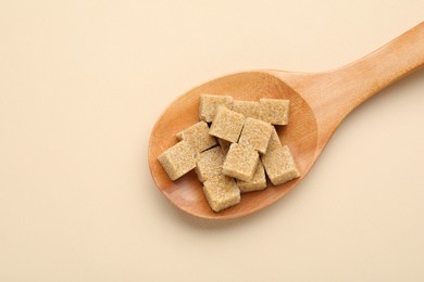 Photo of Brown sugar cubes in wooden spoon on beige background, top view