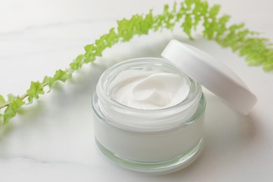 Photo of Face cream in glass jar and green plant on white marble table