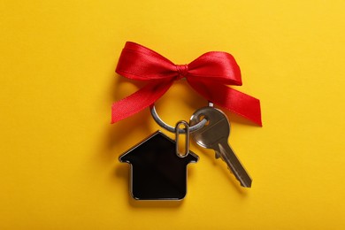 Key with trinket in shape of house and red bow on yellow background, top view. Housewarming party
