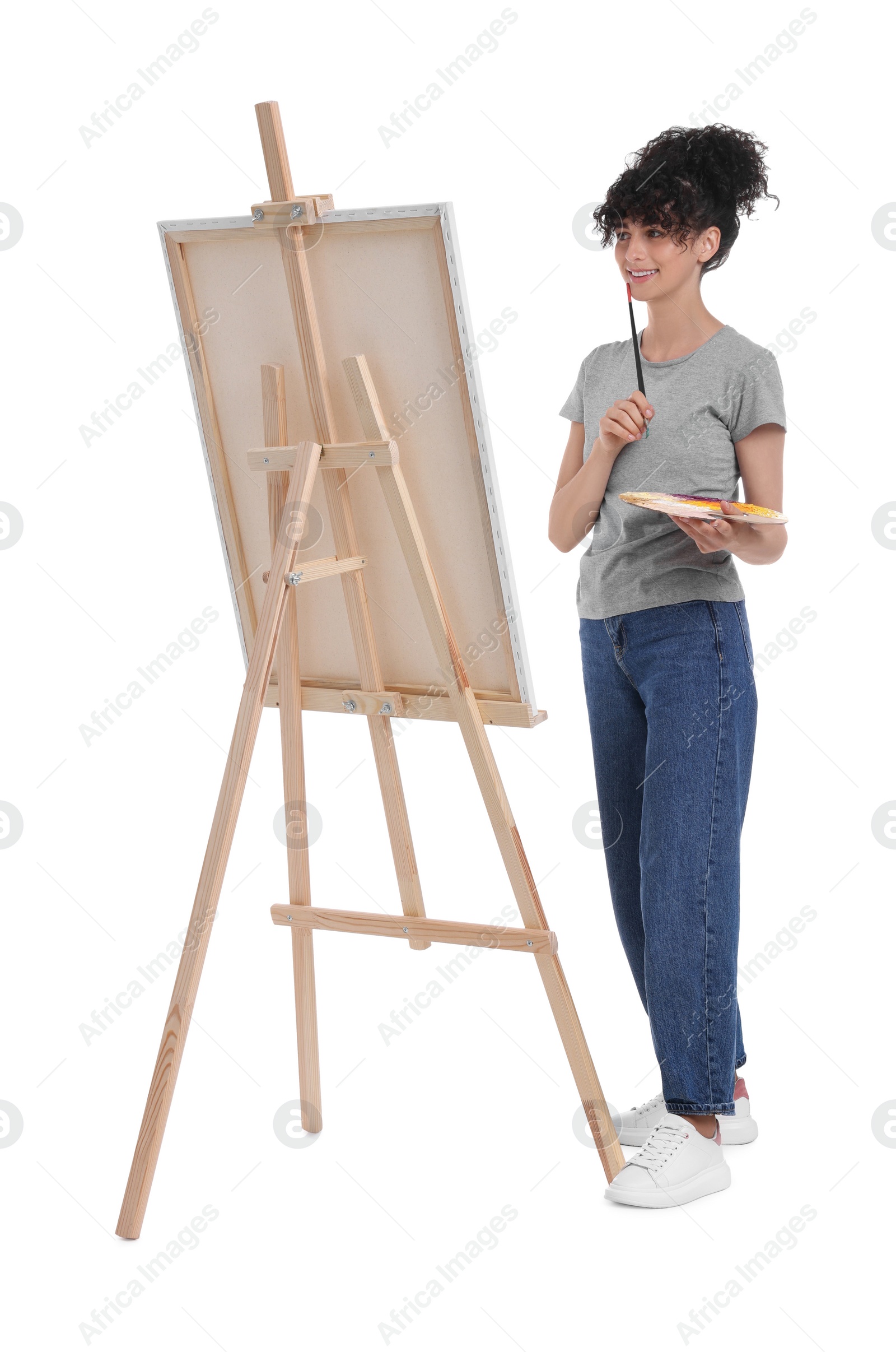 Photo of Young woman holding brush near easel with canvas against white background