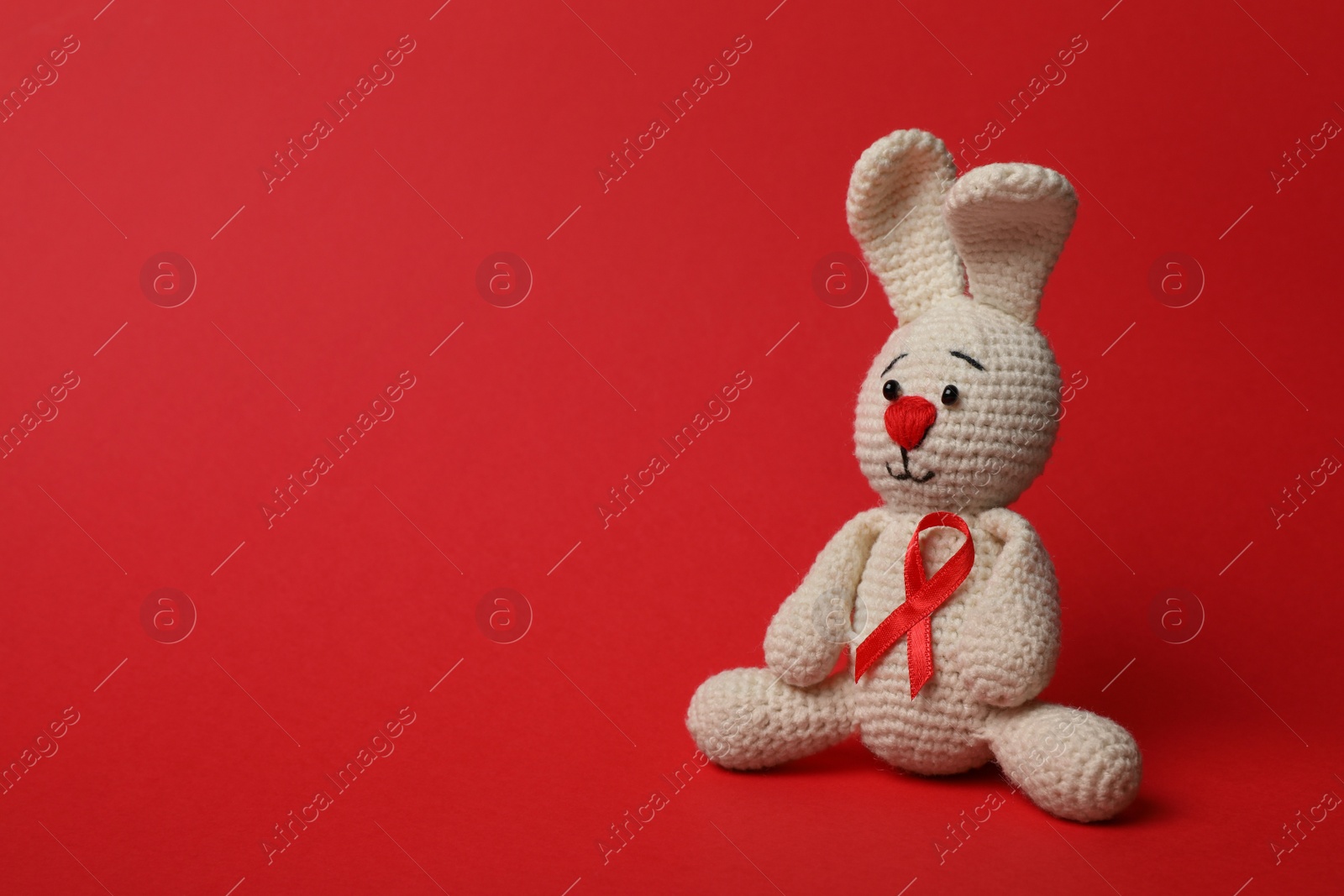 Photo of Cute knitted toy bunny with ribbon on red background, space for text. AIDS disease awareness